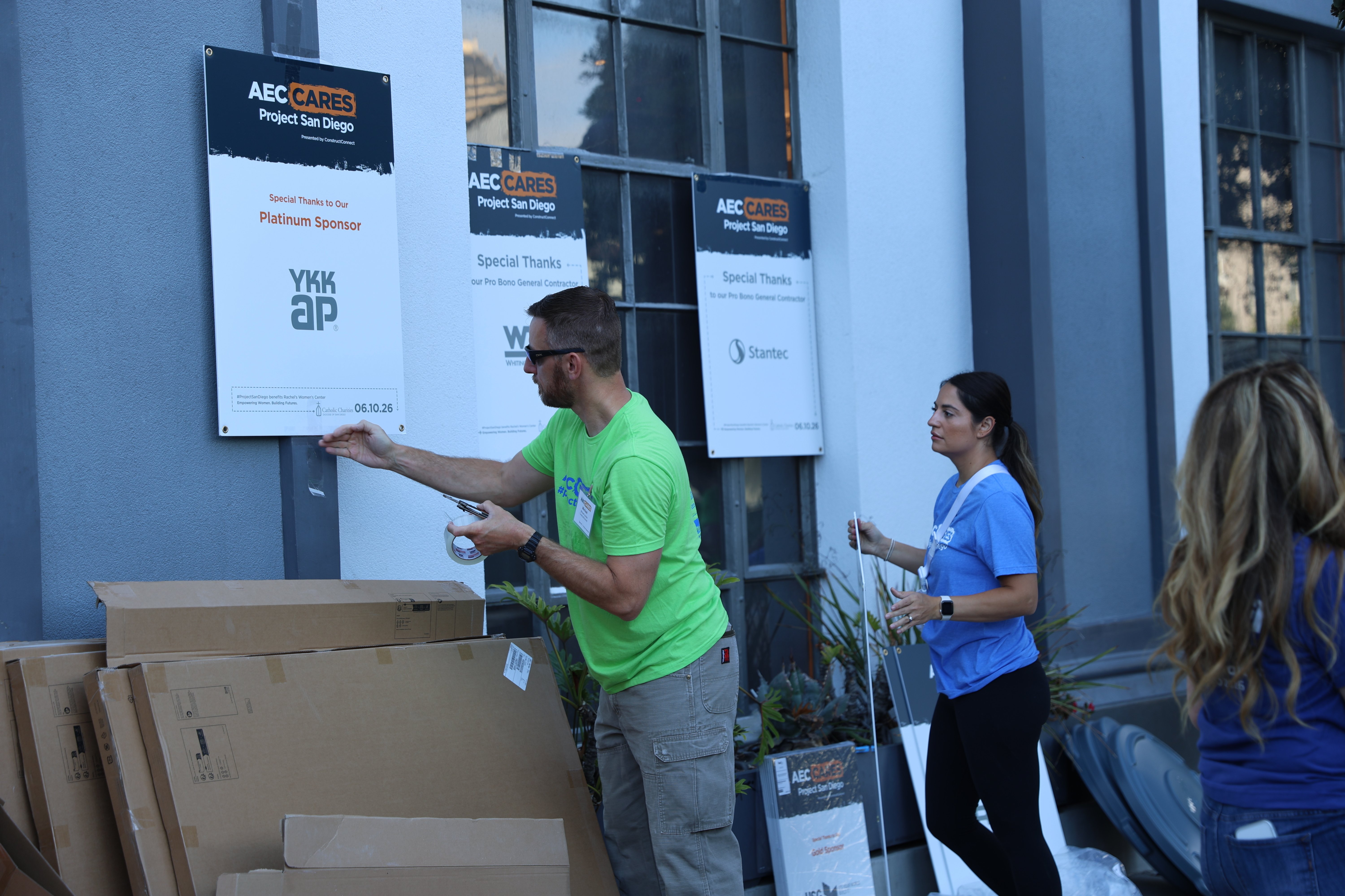 Volunteers hang AEC Cares Project San Diego sponsor signage outside a building with boxes nearby. 