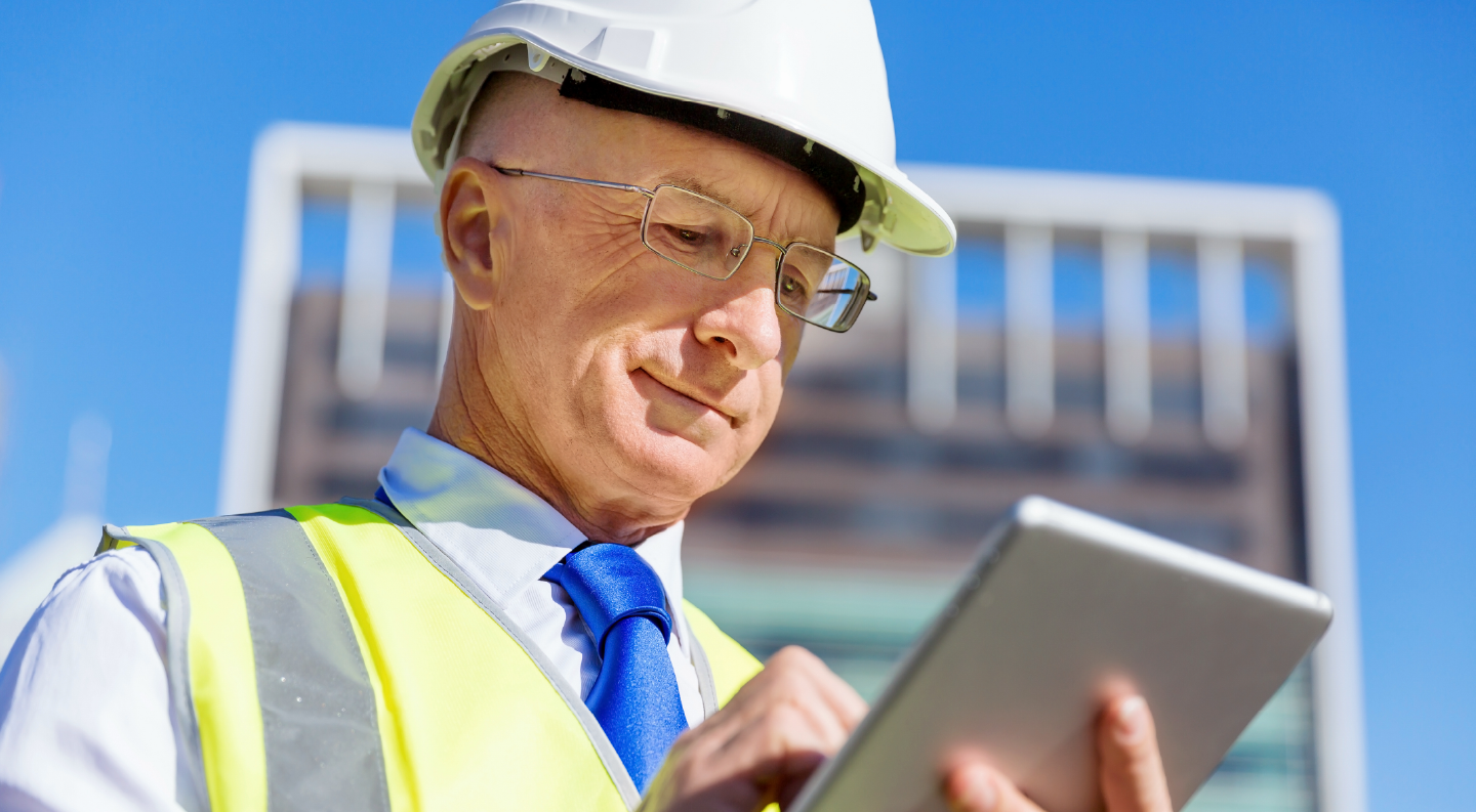 Construction worker with a white hard hat and yellow vest standing on a job site looking at a tablet screen