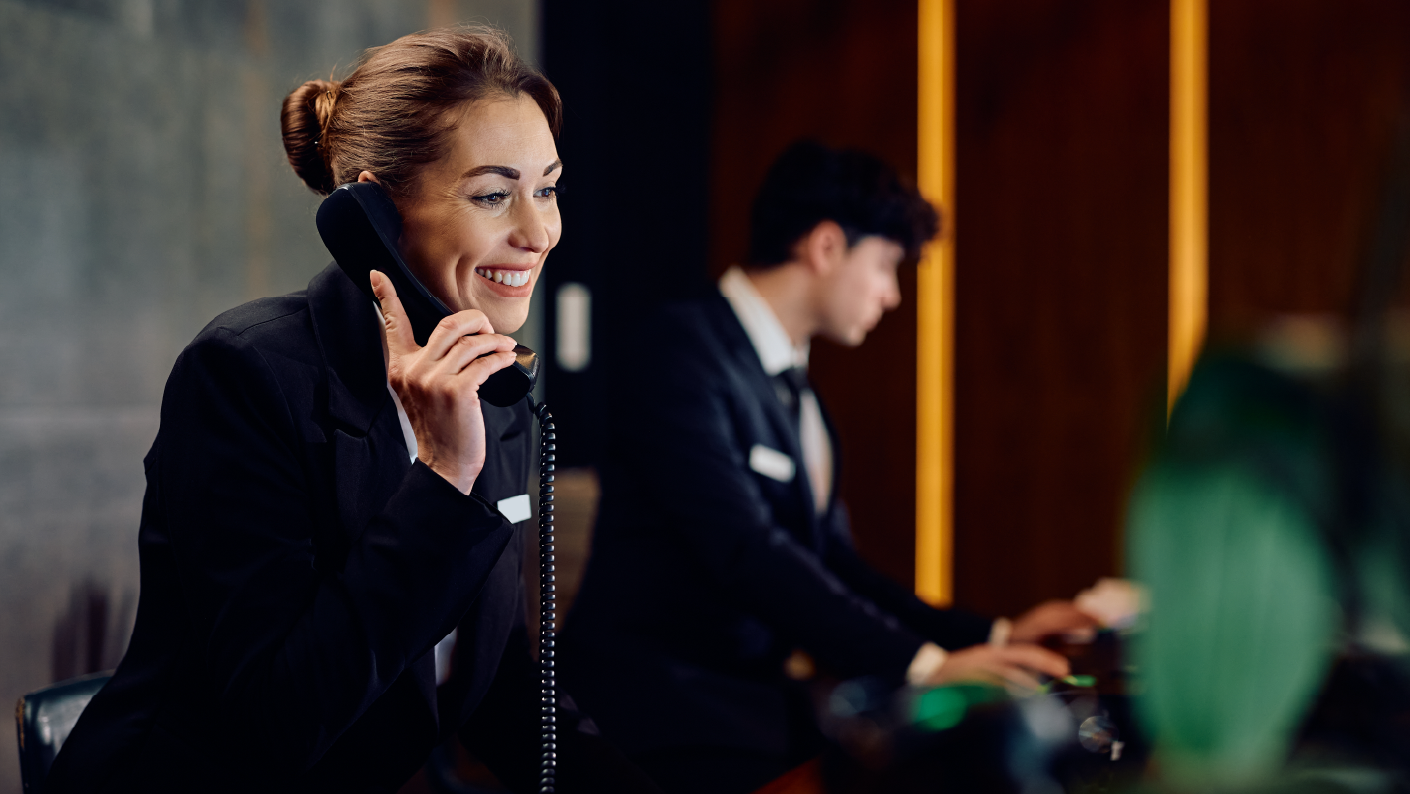 Hotel receptionist speaking on the phone with a colleague working on a computer in the background