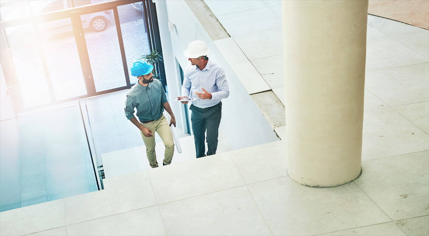 Two construction professionals wearing safety helmets walking up stairs inside a building, discussing plans.