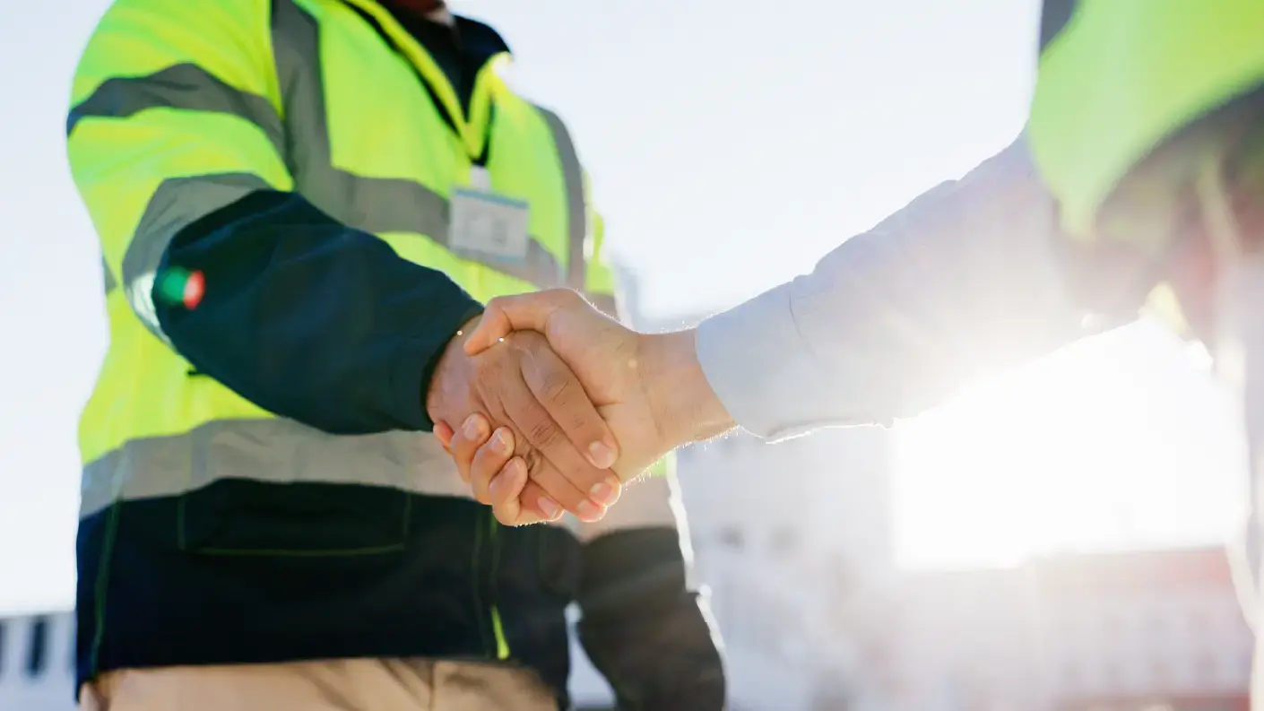 Two people shaking hands outdoors, with one person wearing a high-visibility yellow safety vest with reflective stripes and an ID badge